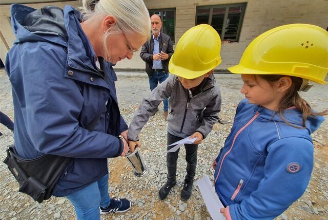 Zwei Grundschulkinder mit gelbem Bauarbeiterhelm befüllen eine Edelstahldose. Sie soll als Zeitkapsel in den Grundstein eines Erweiterungsbaus versenkt werden, der der Massener Schillerschule inzwischen mehr Platz für Ganztagsangebote gibt.
