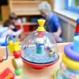 Spielgeräte im Vordergrund und eine Erzieherin und Kinder im Hintergrund