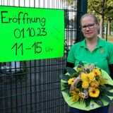 Ein hellgrünes Schild auf dem Steht „Eröffnung 1. Oktober 23. 11 bis 15 Uhr. Daneben eine blonde Frau mit Zopf in einem grünen Shirt und mit einem Blumenstrauß in der Hand.