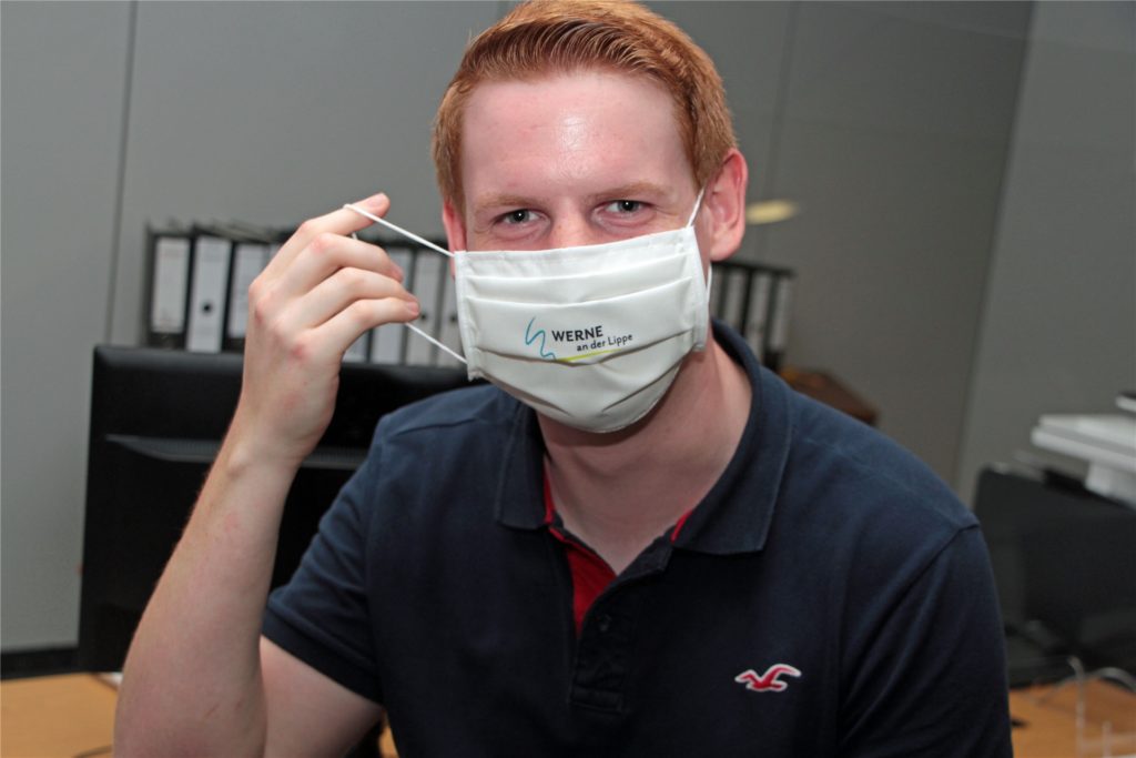 title attribut Verwaltungs-Auszubildender Robin Nolting zeigt die Stoffmaske mit dem Stadt-Logo, die alle Wahlhelfer als Dankeschön erhalten sollen.