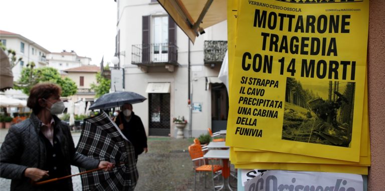 Menschen stehen vor einem Zeitungskiosk, an dem eine Zeitung mit der Schlagzeile „Mottarone-Tragödie mit 14 Toten“ ausgehängt ist. Beim Absturz einer Gondel einer Seilbahn am norditalienischen Lago Maggiore am 23.05.2021 haben mindestens 14 Menschen ihr Leben verloren.