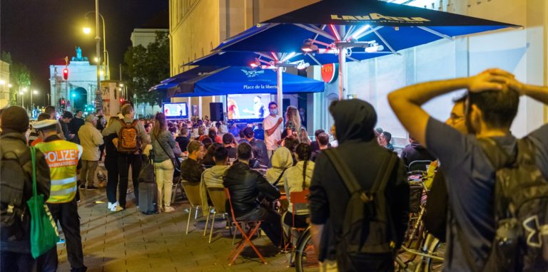 Besucher einer Gaststätte in einer Innenstadt schauen sich auf großen Fernsehschirmen ein Fußballspiel an.
