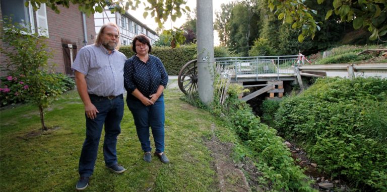 Martin Gockeln und Melanie Skeib-Gockeln vor ihrem Haus in Neimen. Die Brücke im Hintergrund ist die einzige Zufahrt zu ihrem Grundstück – und momentan voll gesperrt. Ein Auto war in die Brücke geschwemmt worden, es der Starkregen Anfang Juli über Fröndenberg hereingebrochen war. Aber auch der Keller der Familie wurde in Mitleidenschaft gezogen.