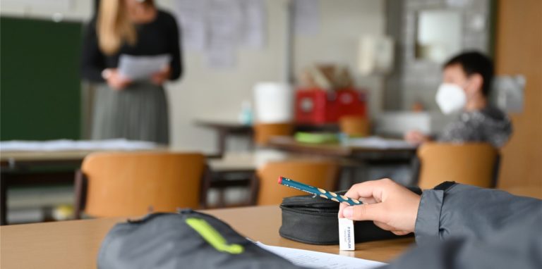 Eine Lehrerin steht mit Maske vor einer Klasse von Schulkindern. Die Kinder tragen ebenfalls eine Maske.