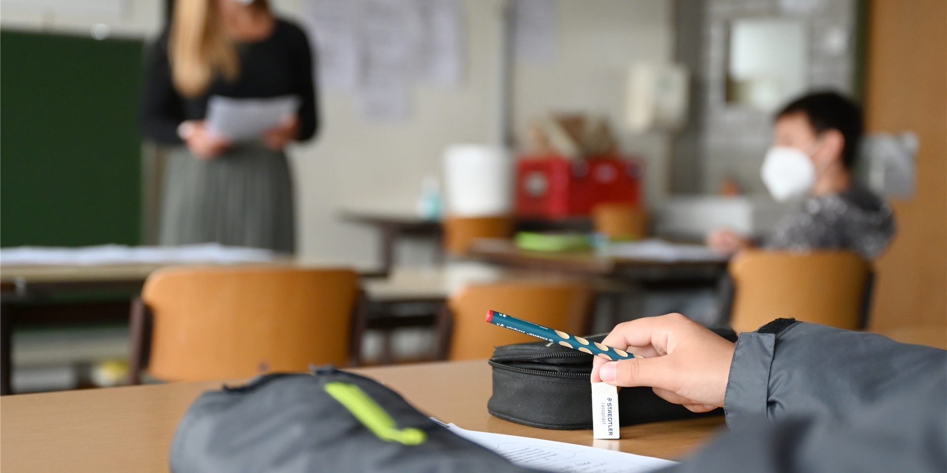 Eine Lehrerin steht mit Maske vor einer Klasse von Schulkindern. Die Kinder tragen ebenfalls eine Maske.