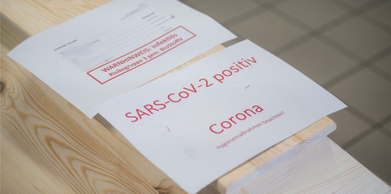 Ein Holzsarg, darauf ein weißes Blatt Papier mit roter Schrift: „SARS-CoV-2 positiv. Corona. Hygienemaßnahmen beachten!“