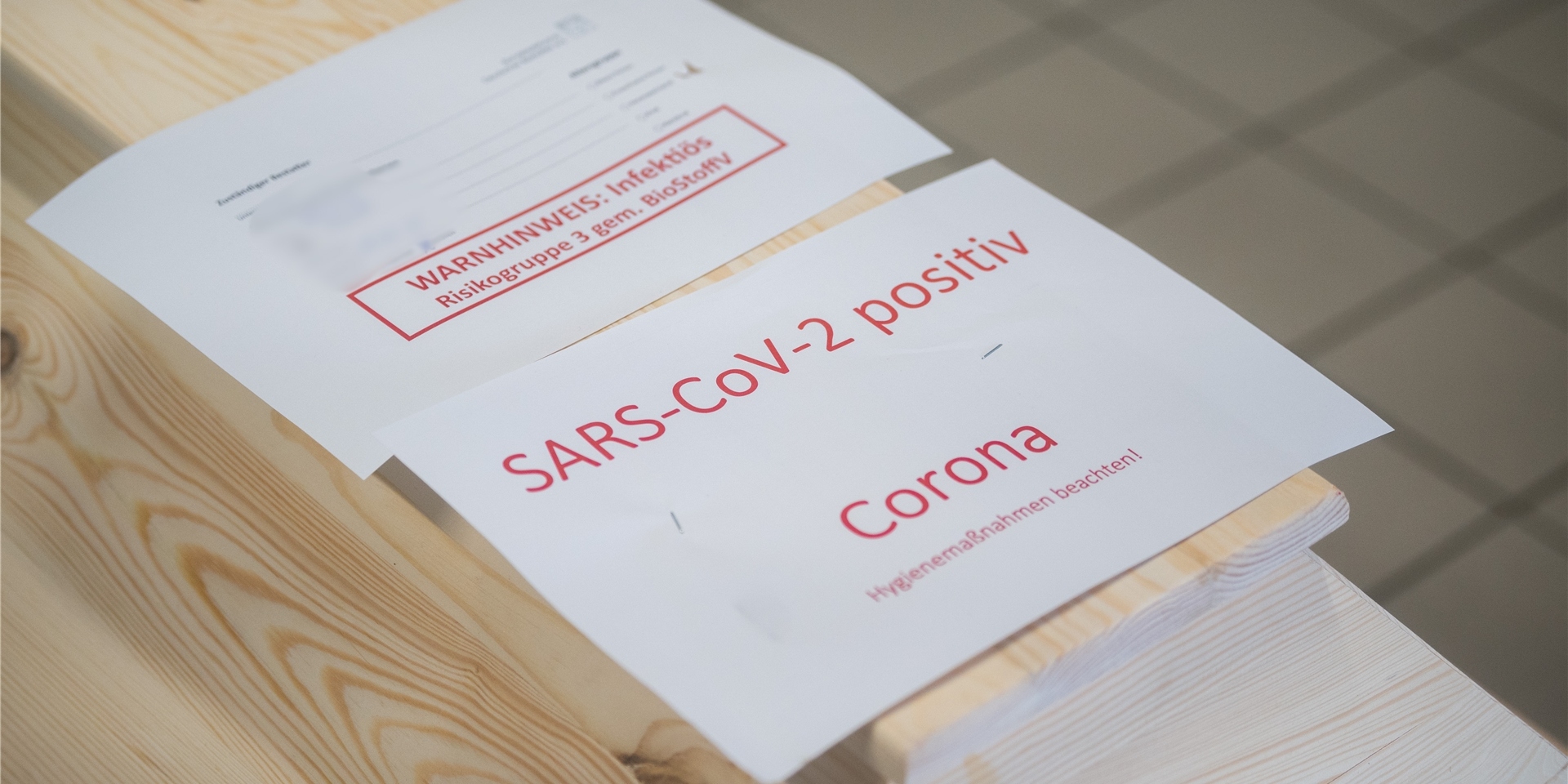 Ein Holzsarg, darauf ein weißes Blatt Papier mit roter Schrift: „SARS-CoV-2 positiv. Corona. Hygienemaßnahmen beachten!“