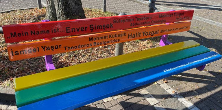 Auf dem Wilhelmplatz sind die Bänke zurück – Initiativen haben sie als Zeichen für Vielfalt bunt gestrichen. Auf einer der Bänke stehen die Namen der zehn Menschen, die von der rechtsextremen Terrorgruppe NSU ermordet worden sind.