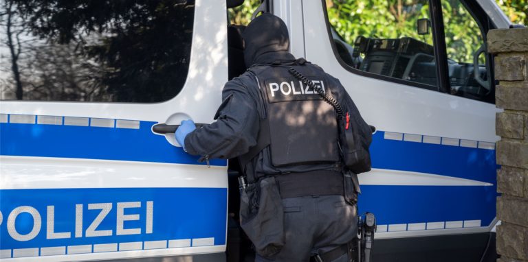 Ein Polizist steht im Rahmen einer Razzia an einem Polizeifahrzeug.