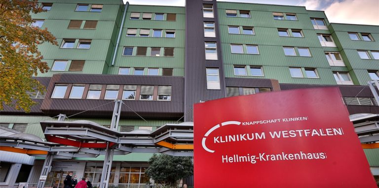 Auf dem Bild erkennt man das Hellmig-Krankenhaus in Kamen. Das Gebäude ist grün und davor steht ein rotes Schild mit dem Namen des Klinikums.