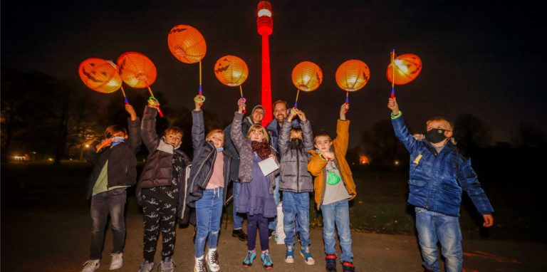 Sieben Kinder und zwei Erwachsenen stehen mit in die Luft gestreckten Kürbis-Laternen auf einem Weg im Westfalenpark. Im Hintergrund wird der Florianturm rot angestrahlt.