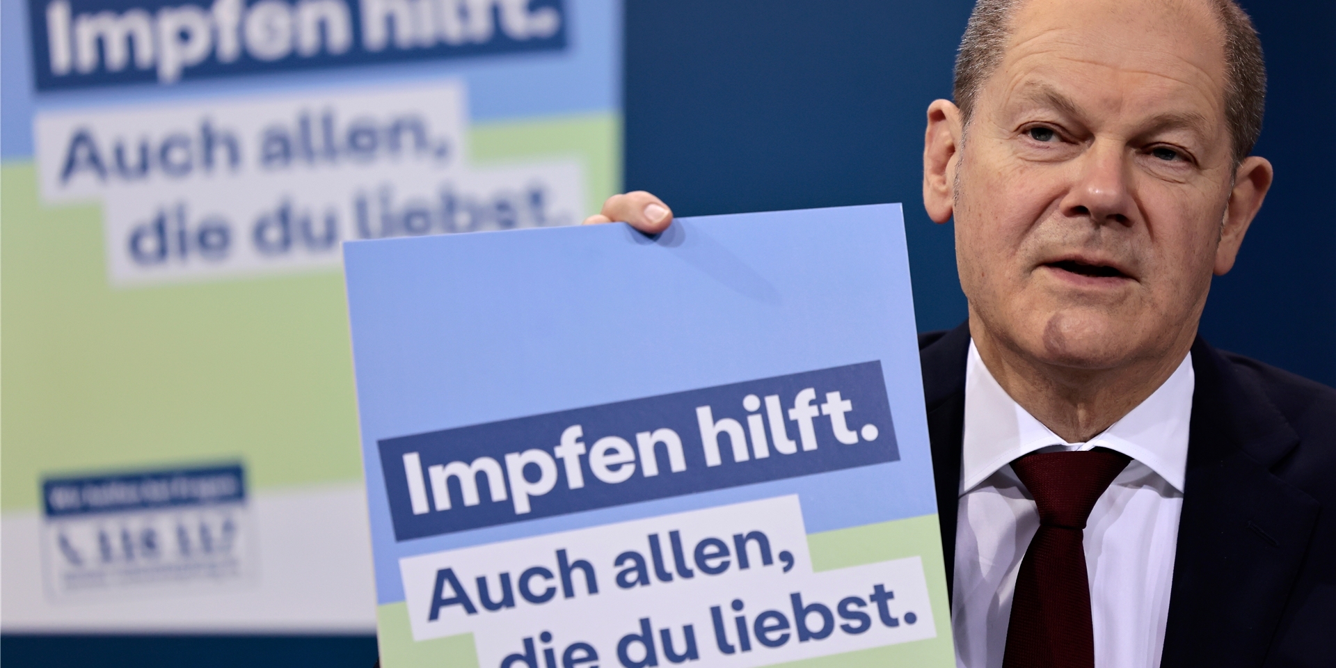 Bundeskanzler Olaf Scholz (SPD) präsentiert ein Plakat für eine neue Impfkampagne während einer Pressekonferenz nach einem Treffen im Kanzleramt. Bundeskanzler Olaf Scholz traf sich mit den Ministerpräsidenten der 16 Bundesländer, um über die Coronavirus-Situation und die Maßnahmen zur Bekämpfung des Virus zu sprechen. Auf dem Plakat steht: „Impfen hilft. Sie hilft auch allen, die man liebt.“