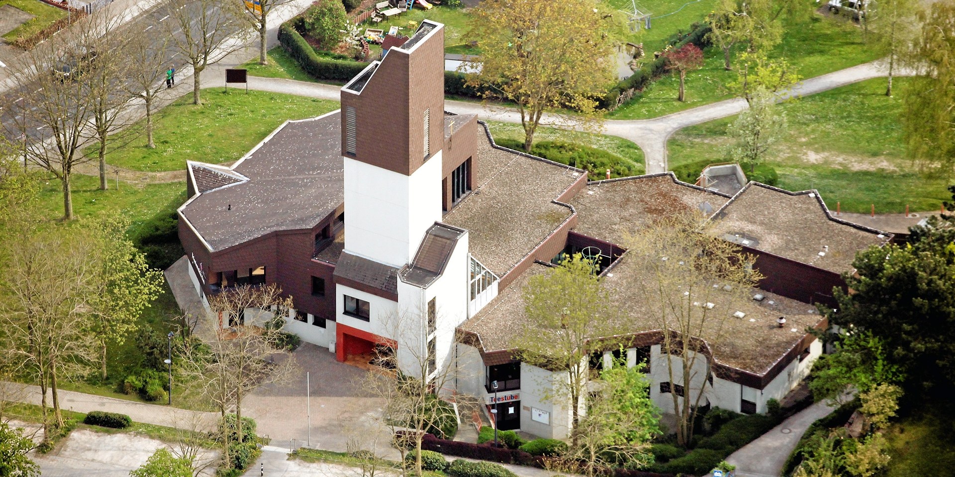 Luftbild einer Kirche mit Parkplätzen, Wiese, Bäumen und Häusern drumherum.