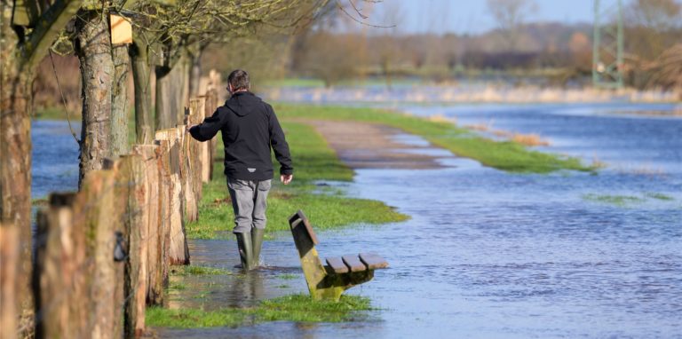 Ein Mann watet mit Gummistiefeln durch das Wasser der über die Ufer getretenen Pinnau. Noch sind viele Felder und Wiesen überschwemmt und nicht alle Straßen wieder trocken. Hochwasser-Situationen könnten durch den Klimawandel öfter auftreten.