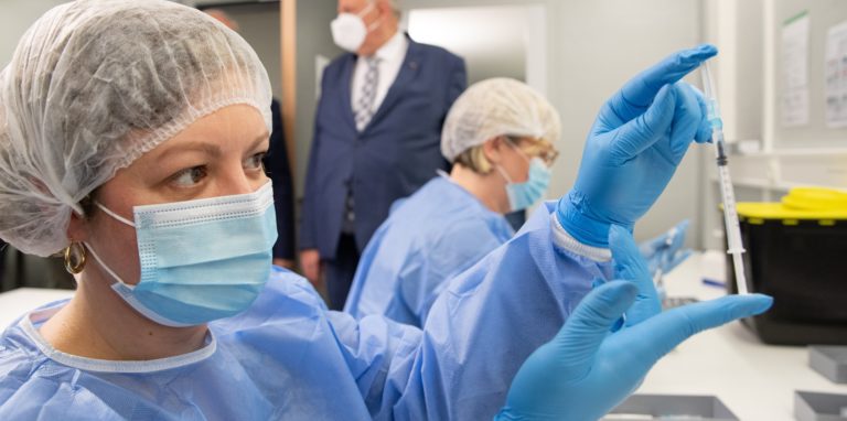 Eine Pharmazeutisch-technische Assistentin (PTA) hält in der Apotheke vom Impfzentrum eine Spritze mit Impfstoff in der Hand. Im Hintergrund steht Karl-Josef Laumann (CDU), Gesundheitsminister von Nordrhein-Westfalen.