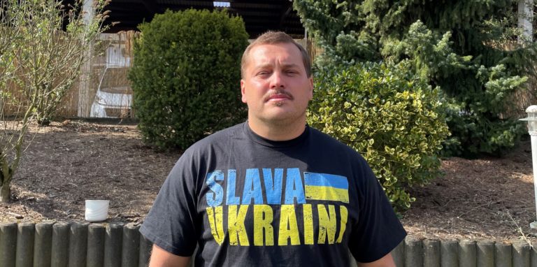 Slava Yatsenko steht im Garten und trägt ein T-Shirt mit dem Aufdruck „Slava Ukraini“.