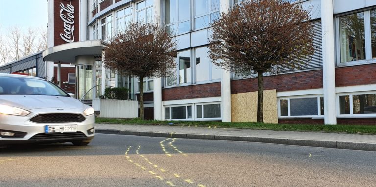 Die gelben Sprühmarkierungen zeigen die Spur des Autos, bevor es schließlich in die Fassade des Coca-Cola-Gebäudes krachte. Das dabei entstandene Loch wurde inzwischen mit einem Holzbrett verschlossen.
