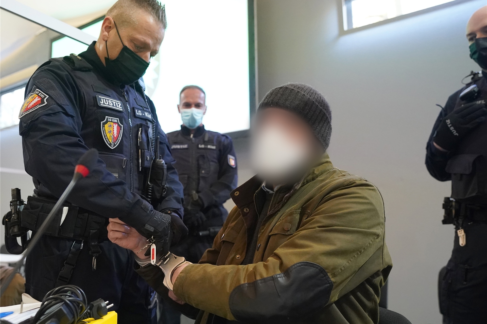Ein Justizbeamter entfernt die Handschellen des Angeklagten (2.v.r) im Sitzungssaal der Außenstelle des Landgerichts zu Beginn des Prozesstages wegen eines Dreifachmordes.