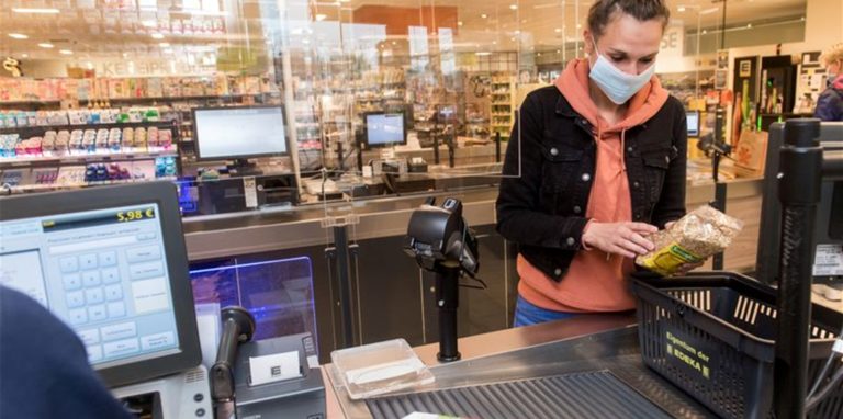 Eine Frau steht an der Kasse im Supermarkt und packt ihre Einkäufe ein. Sie trägt Maske.