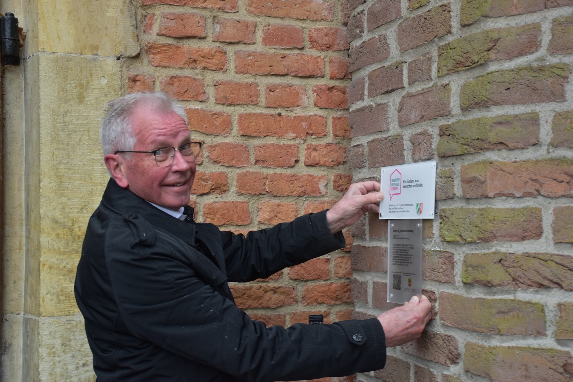 Hubertus Erfmann vom Kirchenvorstand platzierte neue Schilder an der St.-Anna-Kirche. Diese informieren über die Kapelle und weisen auf die Fördermittel für die Beleuchtung hin.