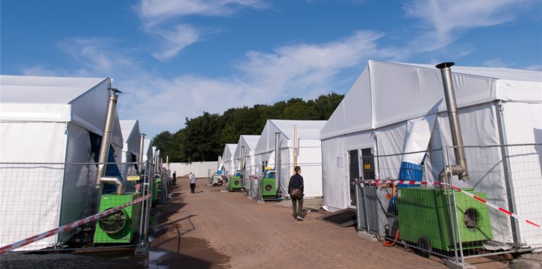 Journalisten und Helfer gehen am 26.08.2015 zwischen den Zelten einer Flüchtlingsunterkunft