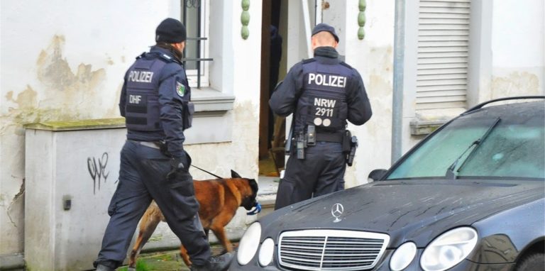 Polizisten und ein Spürhund stehen vor einem Haus.