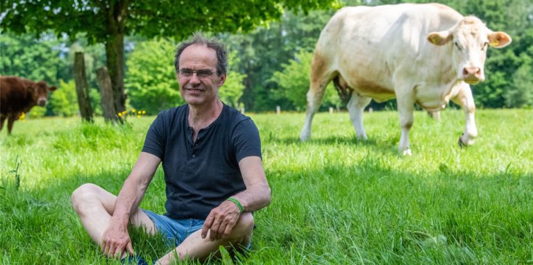 Ulf Allhoff-Cramer, Landwirt, sitzt auf einer Weide neben einer von seinen Kühen. Der Bio-Bauer macht den Autokonzern VW mitverantwortlich für Schäden an seinem Hof. Er will zusammen mit Greenpeace vor Gericht erreichen, dass VW bis 2030 keine Verbrennungsfahrzeuge mehr herstellt.