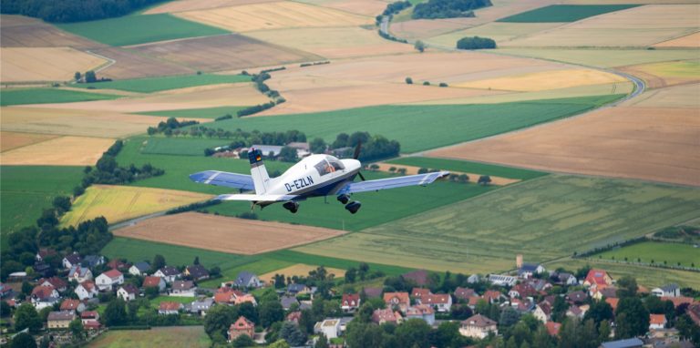 Ein Kleinflugzeug fliegt über eine Siedlung und Felder.