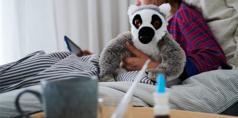 in Fieberthermometer, Nasenspray und eine Tasse Tee stehen vor einem Bett, in dem ein Kind mit Kuscheltier sitzt und auf ein Tablet schaut. Kleine Kinder sind oft krank. Das liegt aber nicht daran, dass ihr Immunsystem nicht gut ist, im Gegenteil: Es ist in Topform und arbeitet auf Hochtouren.