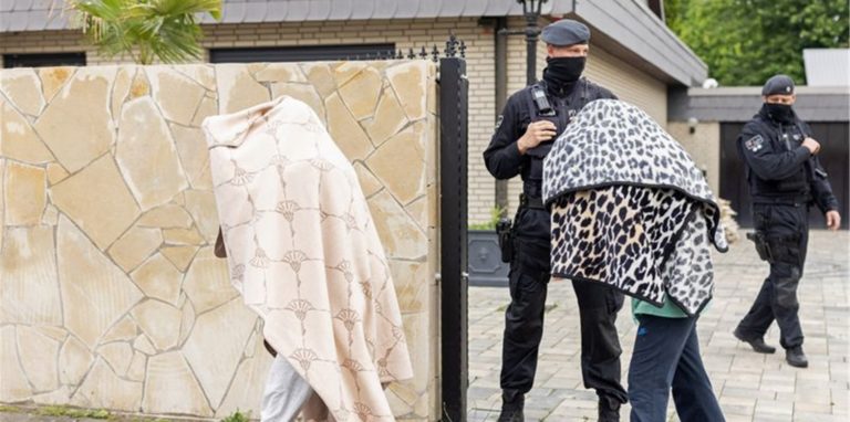 Polizisten stehen vor einer Villa, während zwei Personen mit Decken über dem Körper das Gelände verlassen.