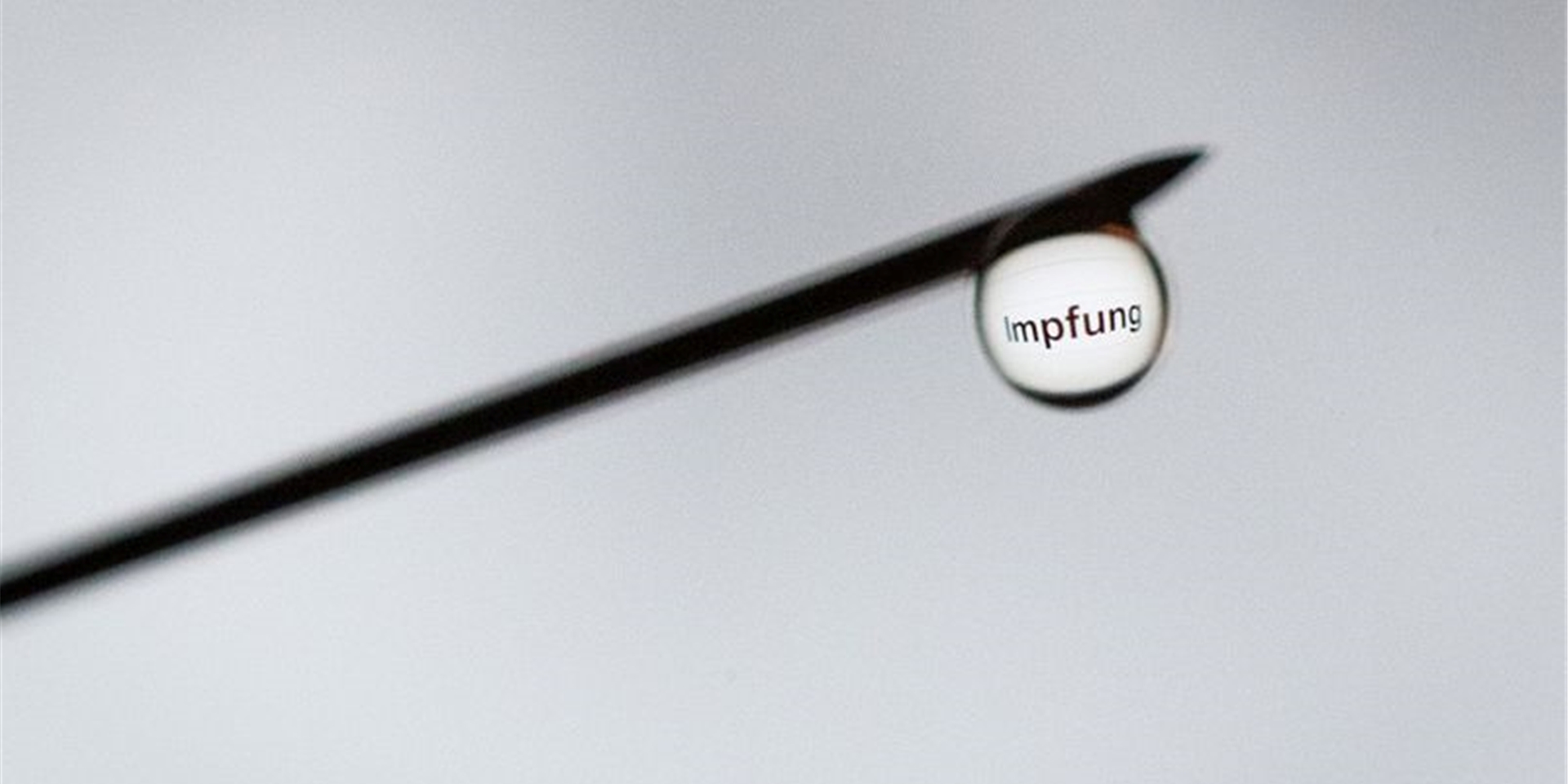 Das Wort „Impfung“ ist in einem Tropfen an der Nadel einer Spritze zu sehen.