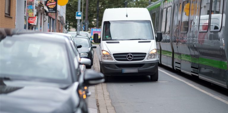 Ein Lieferwagen steht auf einer Straße neben parkenden Autos, daneben steht eine Straßenbahn entlang
