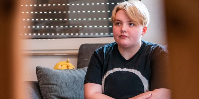 Eine junge Person mit kurzen blonden Haaren sitzt mit verschränkten Armen auf einem Sofa.