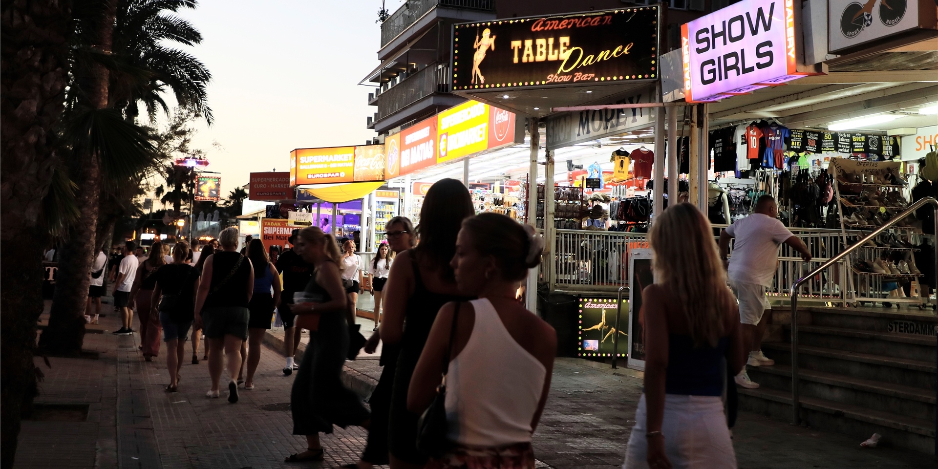 Menschen spazieren auf der Strandpromenade von Arenal auf Mallorca an Show-Bars vorbei.