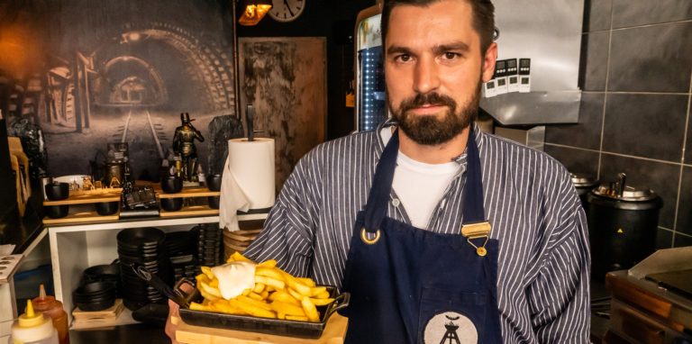 Ein junger Mann mit Bart und Schürze hält eine gusseiserne Schale mit Pommes frites und Mayonnaise in die Kamera.