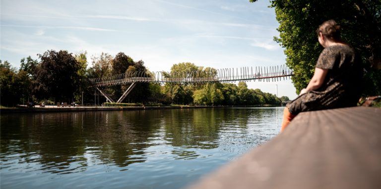 Eine Frau sitzt am Wasser des Rhein-Herne-Kanals und guckt auf die „Slinky Springs to Fame“ Brücke.