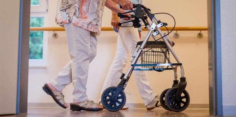 Eine Pflegekraft geht in einem Pflegeheim mit einer älteren Dame über einen Korridor.