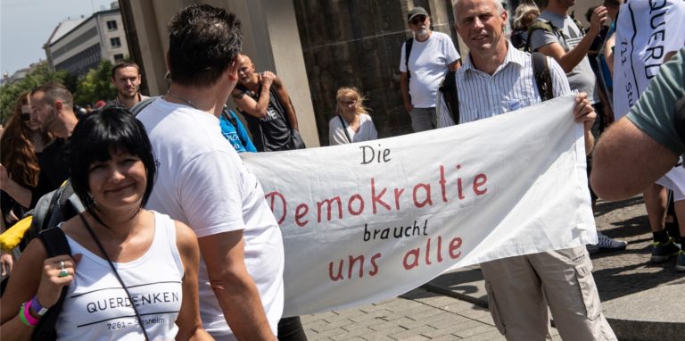 Teilnehmer der Demonstration „Uneingeschränkte Wiederherstellung sämtlicher Grundrechte“ stehen mit einem Transparent mit der Aufschrift „Die Demokratie braucht uns alle“ am Brandenburger Tor.