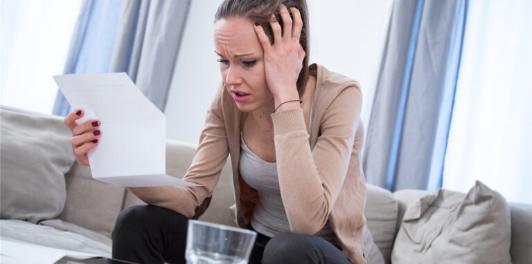 Eine Frau sitzt mit einer Rechnung in der Hand auf der Couch und rauft sich die Haare