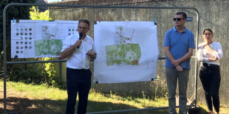 Vor einem Bauzaun, an dem Baupläne befestigt sind, steht Bürgermeister Thomas Orlowski mit Rolf Suhre und einer weiteren Mitarbeiterin des Planungsbüros.