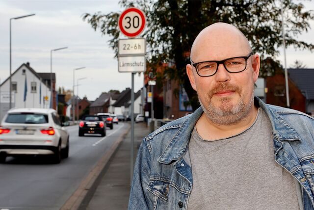Sascha Bähr steht vor seinem Haus an der Lünener Straße. Im Hintergrund stehen Autos in einer Reihe und es ist ein Tempo-30-Schild zu sehen. Bähr selbst trägt eine Glatze, eine Brille und einen grauen Drei-Tage-Bart. Dazu ein graues Shirt und eine helle Jeansjacke.