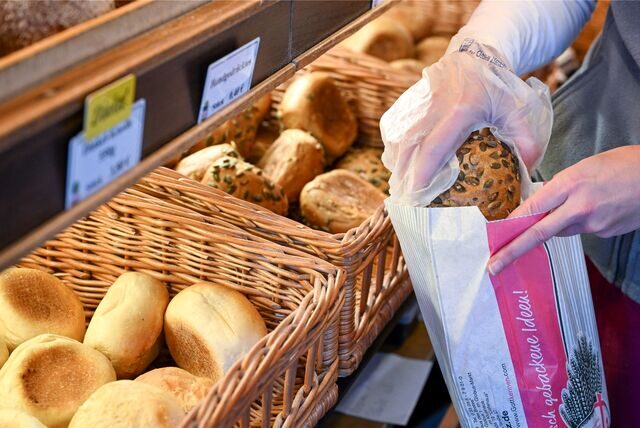 Eine Bäckerei-Verkäuferin nimmt mit einem Handschuh ein Brötchen aus einem Korb und legt es in eine Papiertüte.
