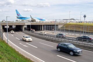 Autos fahren unter einem Teil des Flugfeldes vom Flughafen München hindurch, während ein Flugzeug von TUIfly zum Terminal fährt.