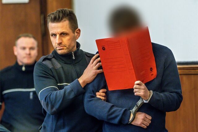 Der Angeklagte wird von einem Beamten zur Anklagebank gebracht und versteckt sich hinter einer Mappe.