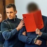Der Angeklagte wird von einem Beamten zur Anklagebank gebracht und versteckt sich hinter einer Mappe.