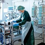 Eine Pflegekraft in einem Krankenzimmer auf der Intensivstation. Die Person trägt einen Schutzanzug, Handschuhe und Maske.