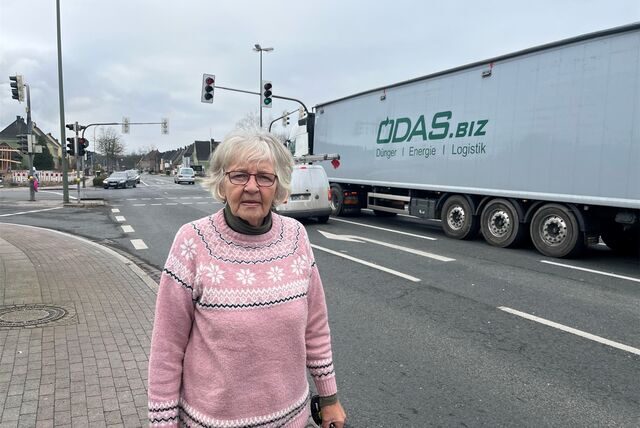 Eine Frau mit rosa Pullover steht an einer Straßenkreuzung. Im Hintergrund ist ein Sattelzug mit Container zu sehen.