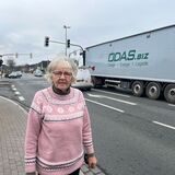 Eine Frau mit rosa Pullover steht an einer Straßenkreuzung. Im Hintergrund ist ein Sattelzug mit Container zu sehen.