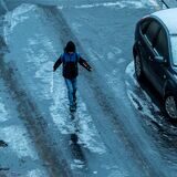 Ein Kind geht vorsichtig über eine vereiste Straße. Es scheint Winter zu sein. Das Kind trägt eine dicke Jacke und eine Mütze. Am Rand der Straße parken Autos. Die Straßenoberfläche sieht spiegelglatt aus.