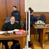 Ein Anwalt und die Beschuldigte sitzen an Tischen im Landgericht Dortmund. Hinter ihnen stehen ein Polizist und eine Polizistin.